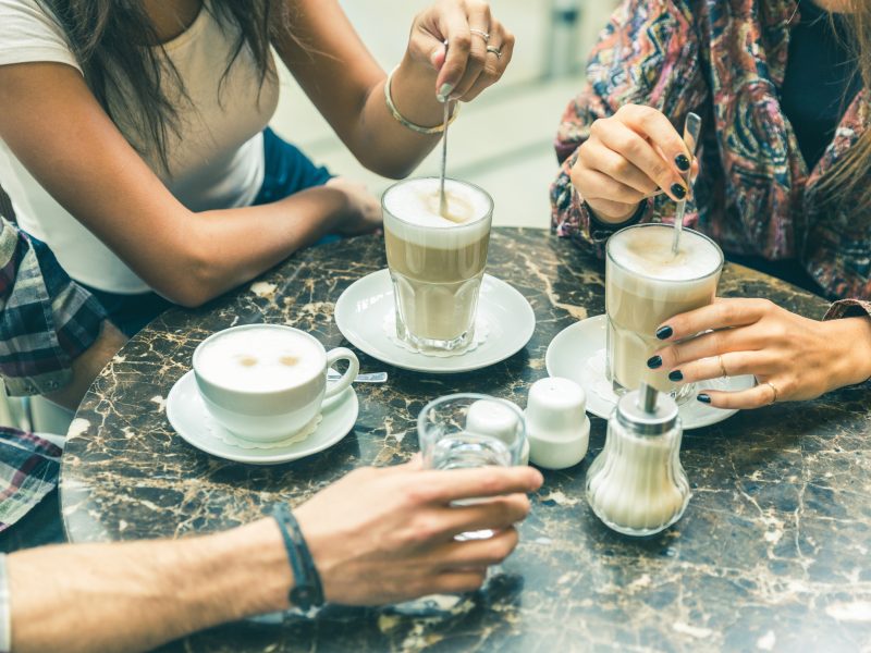 Multiracial group of friends at cafe together. Two women and a man at cafe, focus on glasses and cups, with coffee and cappuccino. Friendship and coffee culture concepts with real people models.